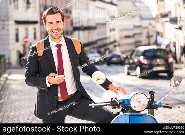 Portrait of happy young businessman with cell phone and motor scooter in the city, Lisbon, Portugal