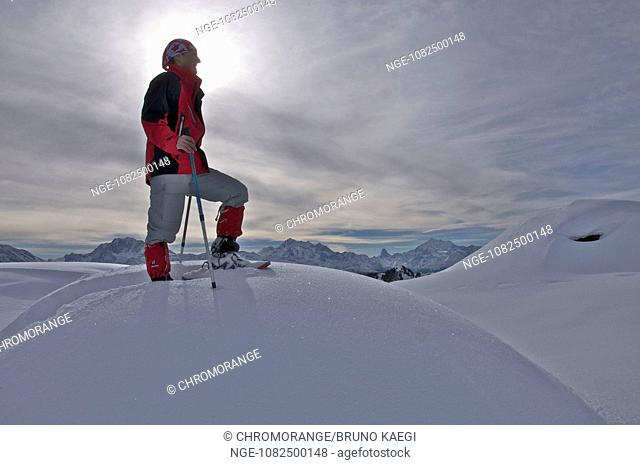 female snowshoer in red windbreaker silhouetted against dramatic sky with snowheavy clouds and sunray