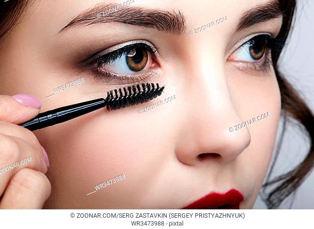 Close up portrait of beautiful young brunette woman face. Female applying mascara to eyelashes by makeup brush