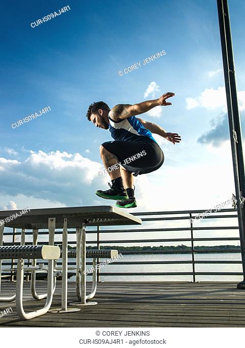 Young man jumping backwards from picnic table on lake pier
