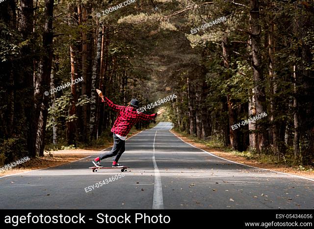A young hipster in a cap and checkered shirt rides on his skateboard on a country road in an autumn forest and makes a stunt slide standing
