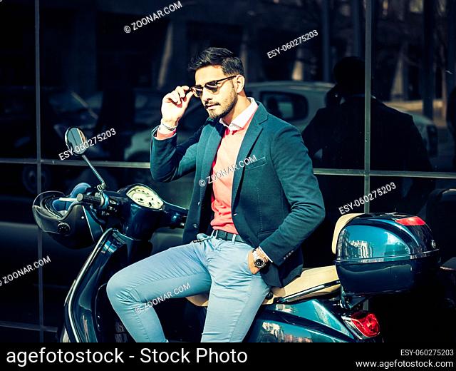 Young stylish man in suit and sunglasses standing by his classic Italian scooter on empty road in sunny city