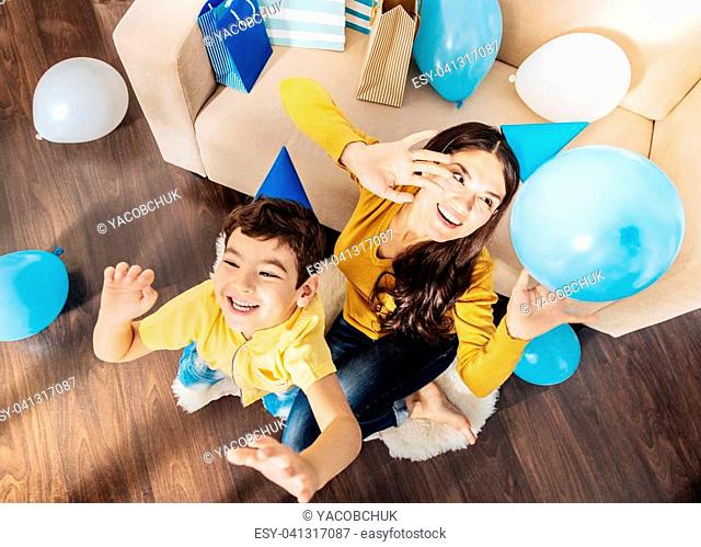 Enjoying themselves. Upbeat little boy and his pleasant mother sitting on the rug near the sofa and playing with balloons while laughing