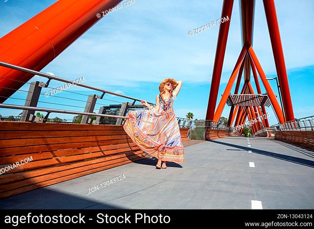 Female wearing a long summer sway dress of vibrant colours and straw hat, walking across the new bridge Yandhai Nepean Crossing