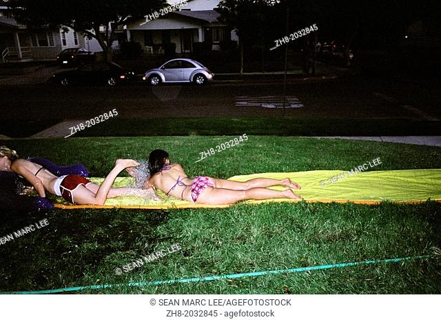 Young women in bikini's slide on a slip and slide type water amusement toy on a lawn at night in Los Angeles