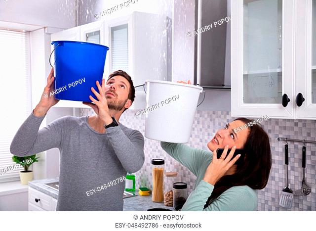 Worried Young Woman Calling Plumber On Cellphone While Man Collecting Leakage Water From Ceiling