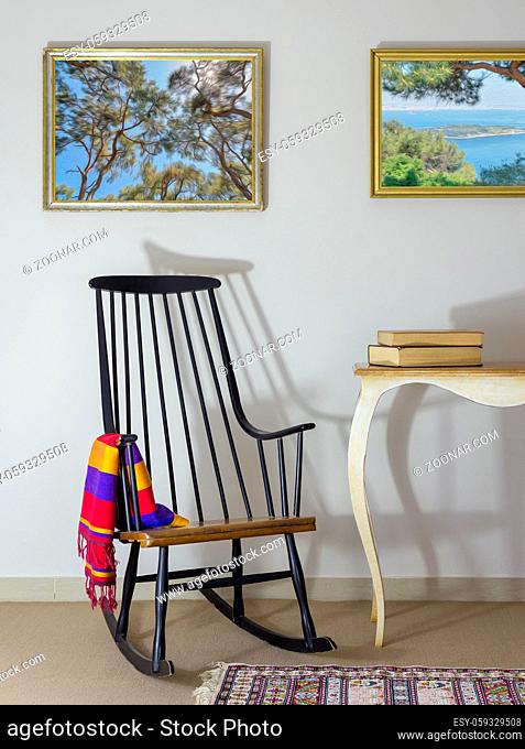 Interior shot of classic rocking chair and two old books on old style vintage table on background of off white wall with two hanged paintings including clipping...