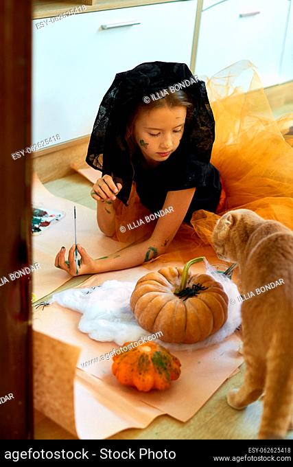 Happy child decorating a pumpkin at home with cat, Little girl drawing face on orange Halloween Jack-O-Lantern pumpkin. Holiday decoration concept