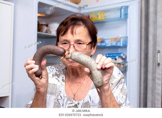 Senior woman holding pork liver sausages while standing in front of the open fridge in the kitchen