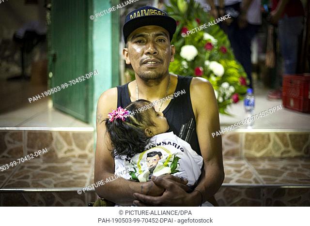 03 May 2019, Mexico, Culiacán: A man is holding a girl with a Malverde T-shirt on her arm and is wearing a cap with the name of Jesus Malverde