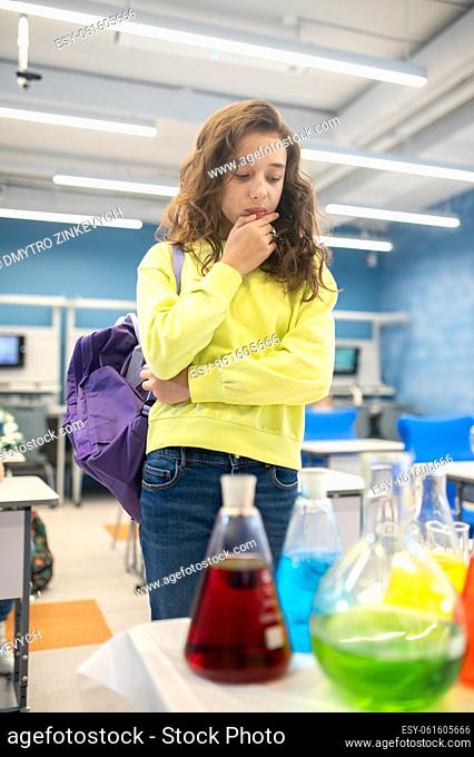 Interesting here. Interested serious girl touching chin examining chemical flasks with liquid standing in lighted classroom at school