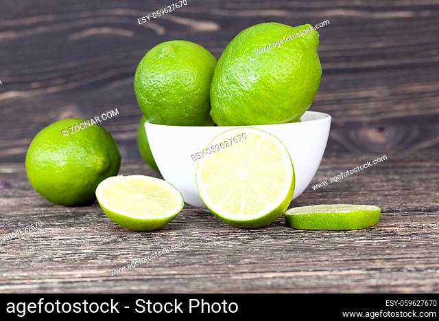 chaotically scattered on the table green ripe sour limes, a black background with a bowl in which lie the fruit, one lime is chopped