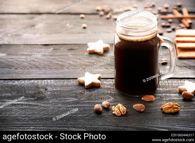 Big glass jar full with hot coffee and surrounded by cinnamon star shaped cookies, walnuts and almonds, on a rustic black wooden table, under the morning light