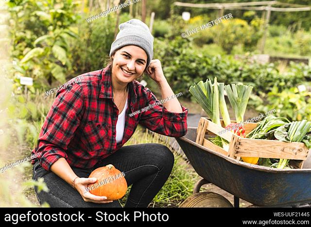 Smiling young woman holding squash by wheelbarrow in vegetable garden