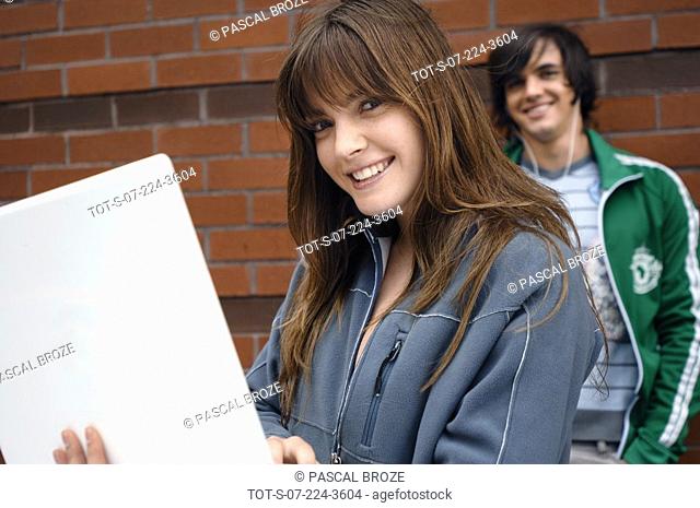 Portrait of a young woman using a laptop with a young man leaning against a brick wall in the background