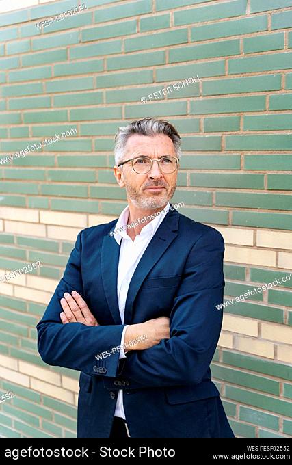 Confident mature businessman with arms crossed standing against wall
