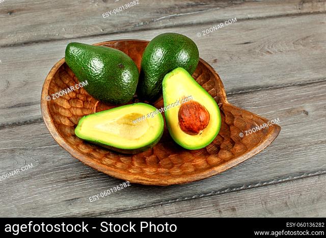 Three avocados, one of them cut to half, seed visible, in carved wooden bowl