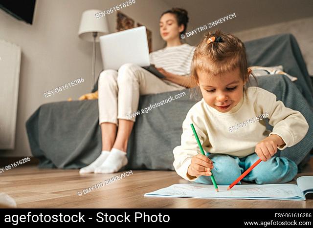 Young woman typing on the laptop while her daughter sketching with pencils in the sketchbook