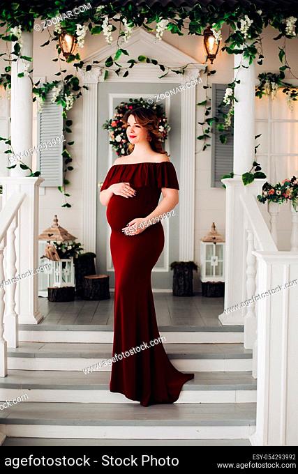 Gorgeous pregnant woman wearing in marsala dress posing, touching her belly. Beautiful future mother standing on steps of white house