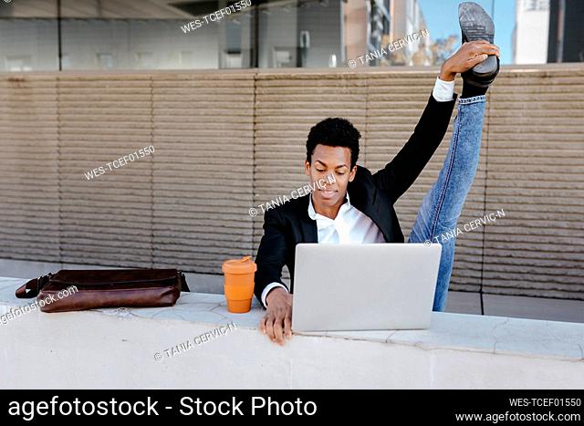 Flexible male entrepreneur using laptop while sitting at retaining wall