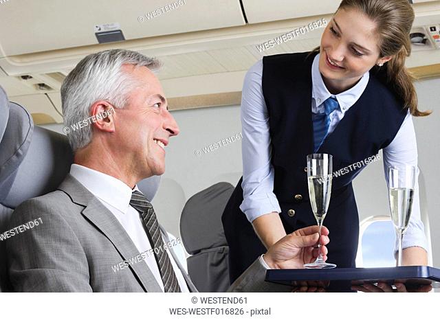 Germany, Bavaria, Munich, Young stewardess serving champagne to senior businessman in business class airplane cabin