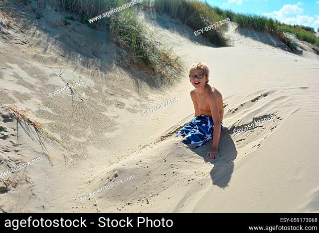 Boy sits in the sand dunes and has his legs dug in the sand, laughs forward
