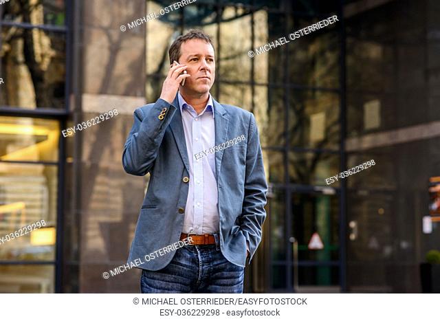 A middle age businessman standing in front of an office building while talking on his phone