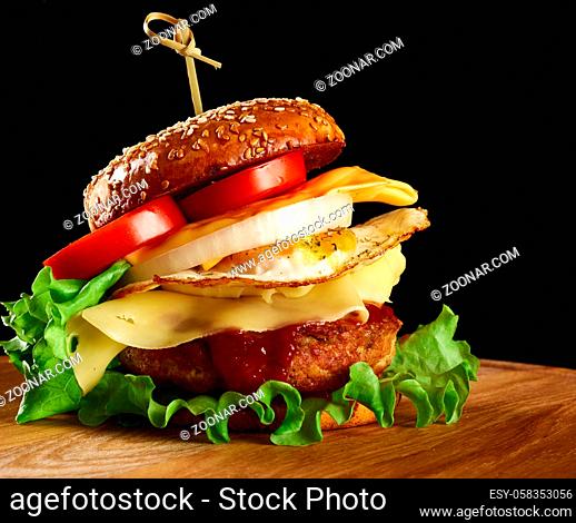 burger with meat cutlet, cheese, fried egg, tomatoes, cucumber slices and green lettuce, fast food on a round wooden board, black background