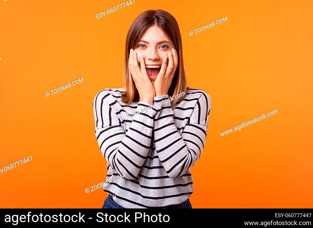 Portrait of shocked young woman with brown hair in long sleeve striped shirt standing, looking with amazement and hiding face in arms, unexpected news