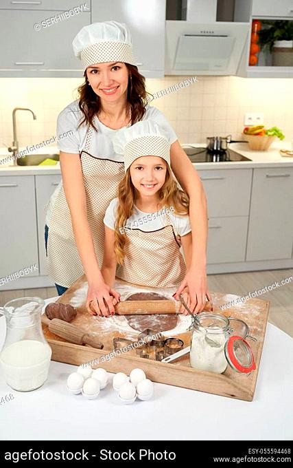Mother and daughter squeezing dough for chocolate cookies on pastry board. Brunette woman and girl in aprons and chef hats holding rolling pin together, smiling