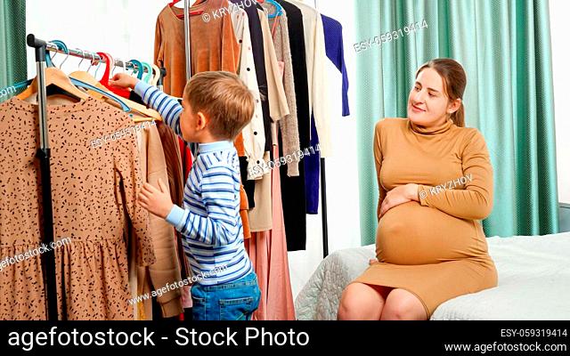 Happy smiling boy taking clothes from clothes rack ad giving it his mother in wardrobe