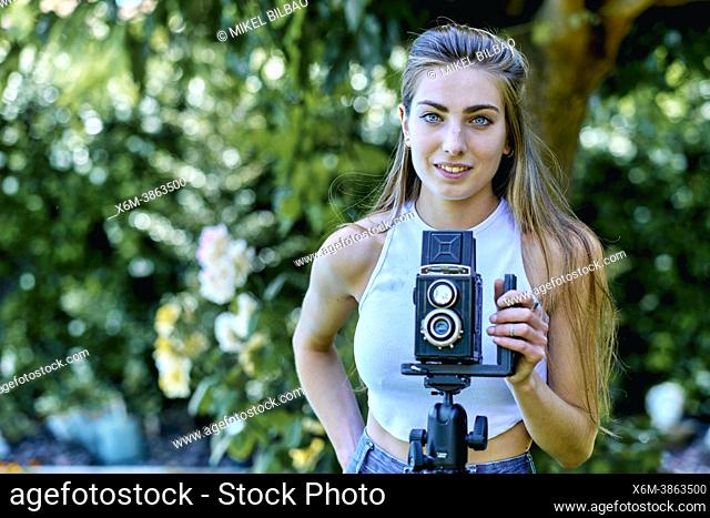 Portrait of a young beautiful caucasian woman in her 20's photographing with an old vintage camera on a tripod outdoor in a garden. Lifestyle concept