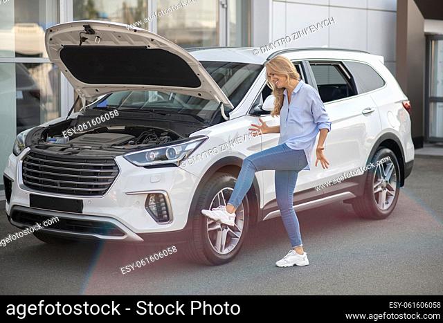 Problem, desperation. Emotional young adult woman gesturing kicking tire of car standing with open hood near building outdoors