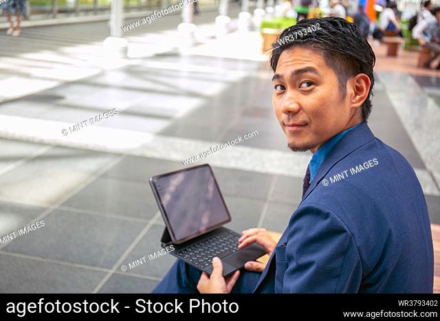 A young businessman in a blue suit in a city downtown area, sitting on a bench using a digital tablet looking over his shoulder