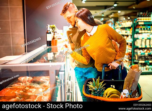 Family couple choosing fresh chilled meat in grocery store. Man and woman with cart buying beverages and products in market, customers shopping food and drinks