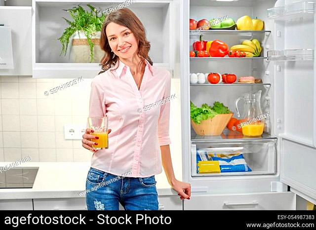 Front view of pretty woman in white shirt and jeans keeping glass of orange juice, looking at camera and posing at kitchen