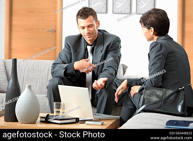 Young businessman explaining to businesswoman at office sitting on sofa talking, looking at laptop computer