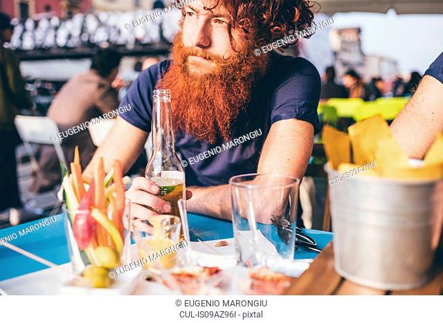 Young male hipster with red hair and beard drinking bottled beer at sidewalk bar