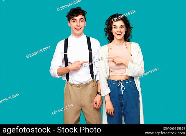 Portrait of happy successful two brunette stylish partner, couple or friends standing and pointing each other and looking at camera with toothy smile