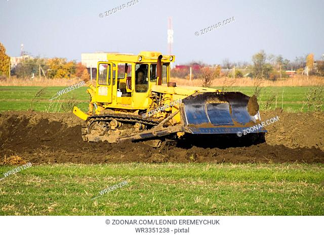 The yellow tractor with attached grederom makes ground leveling. Work on the drainage system in the field