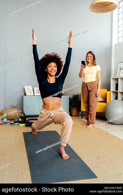 Woman photographing friend exercising with arms raised through smart phone at home