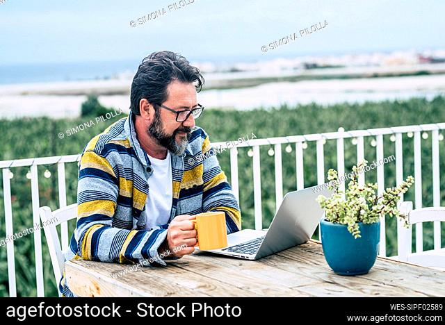 Bearded man with coffee mug using laptop while working on rooftop