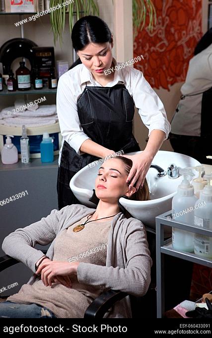 Side view of beautiful hairdresser washing hair to her client lady in hairdressing saloon. Client sitting with her eyes closed