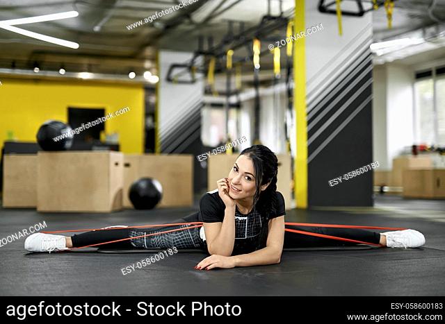 Athletic girl does a twine with a red fitness rubber on the gray mat in the gym. She wears a dark sportswear with white sneakers