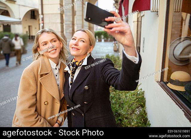 Smiling elegant mature blonde woman photographing herself and a beautiful young lady against the city sights