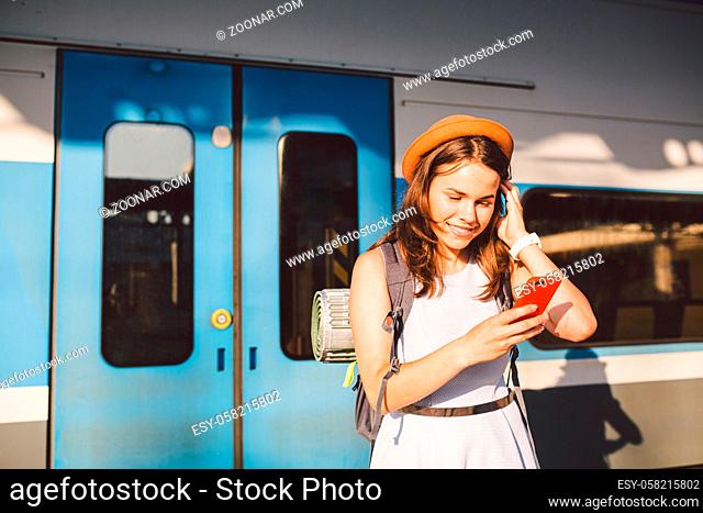 Railroad theme. Beautiful young woman with a backpack uses the phone while standing near the railroad train on the platform. Cheap travel