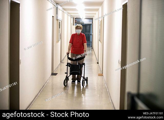 Senior woman wearing mask and walking with wheeled walker in corridor of retirement home