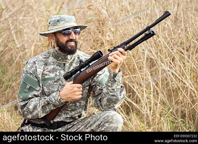 The smiling hunter in military uniform is holding rifle weapon or airgun and squatting in nature