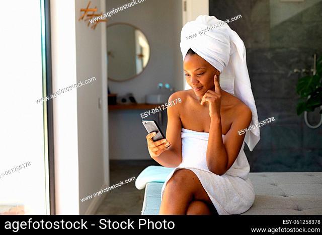 African american woman in a bathrobe using smartphone sitting on the bed at home