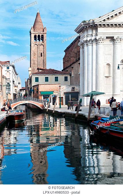 Motorboat and barge moored alongside canal with reflections of Renaissance church and Romanesque bell tower in water, people wandering in piazza